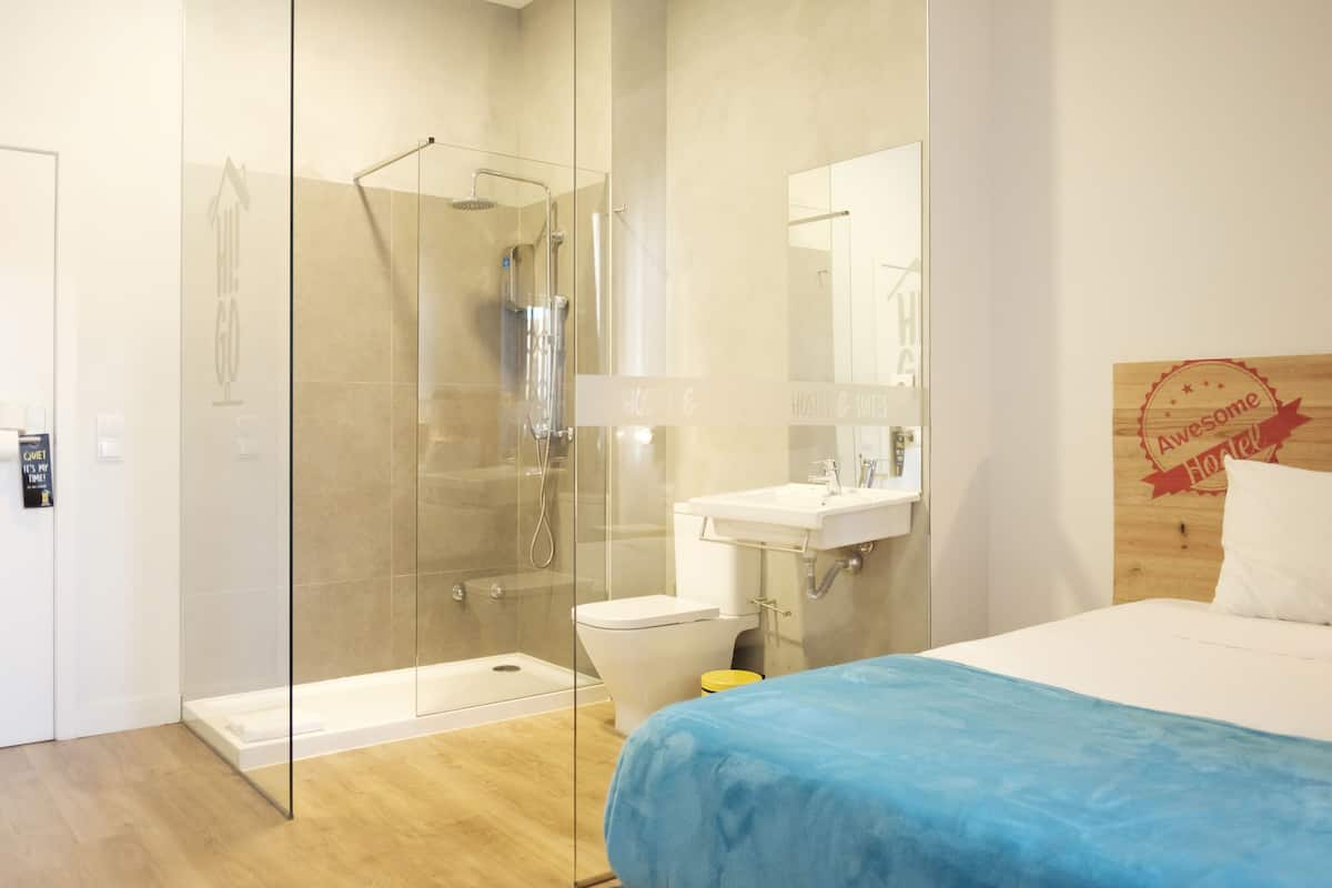 Une chambre d’hôtel moderne dotée d’une douche à parois de verre, de toilettes blanches, d’un lavabo et d’un lit recouvert d’une courtepointe bleue.