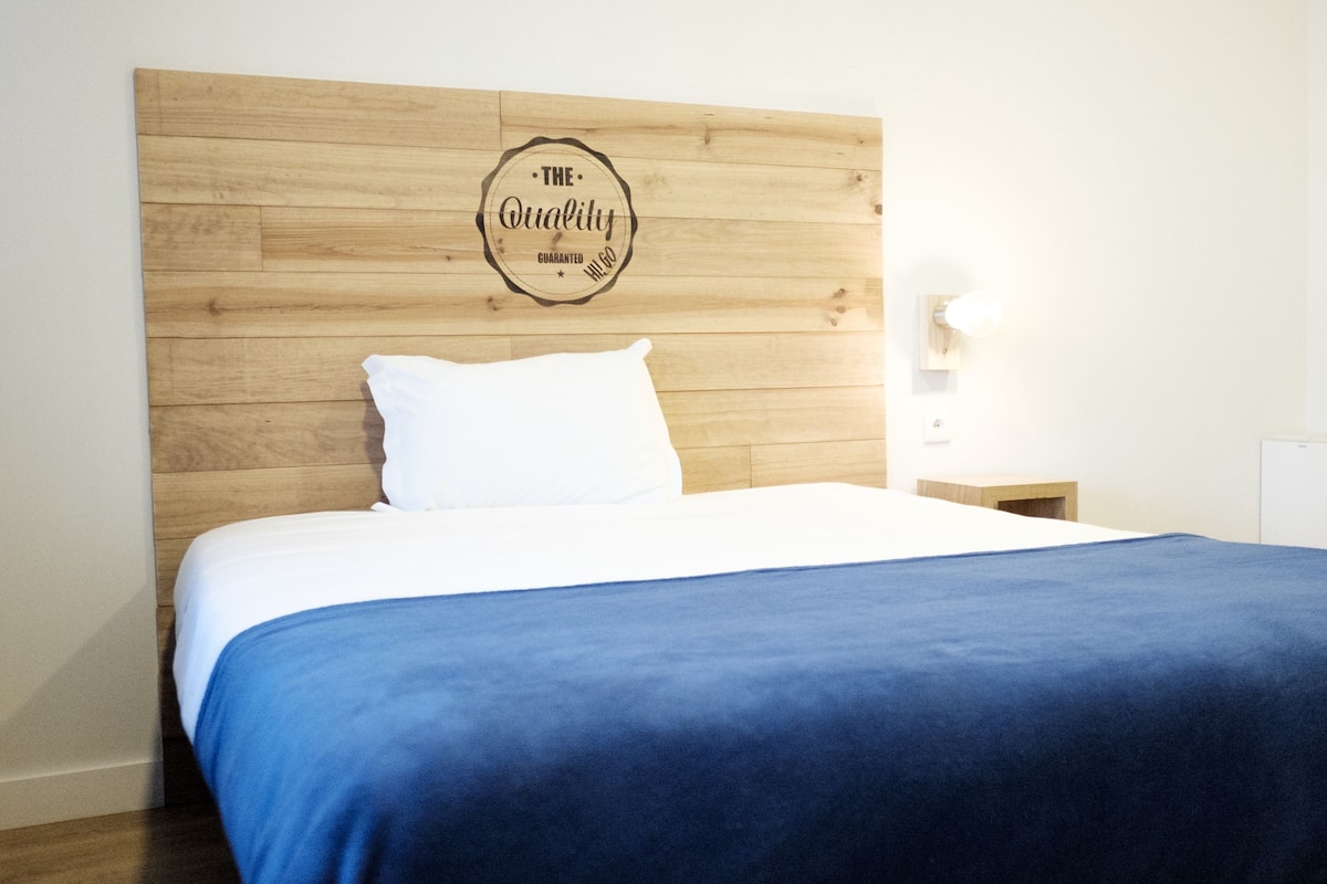 Un lit bien fait, avec une couvre-lit bleue et des oreillers blancs, une tête de lit en bois ornée d’un motif circulaire, et une petite table de chevet avec une lampe.
