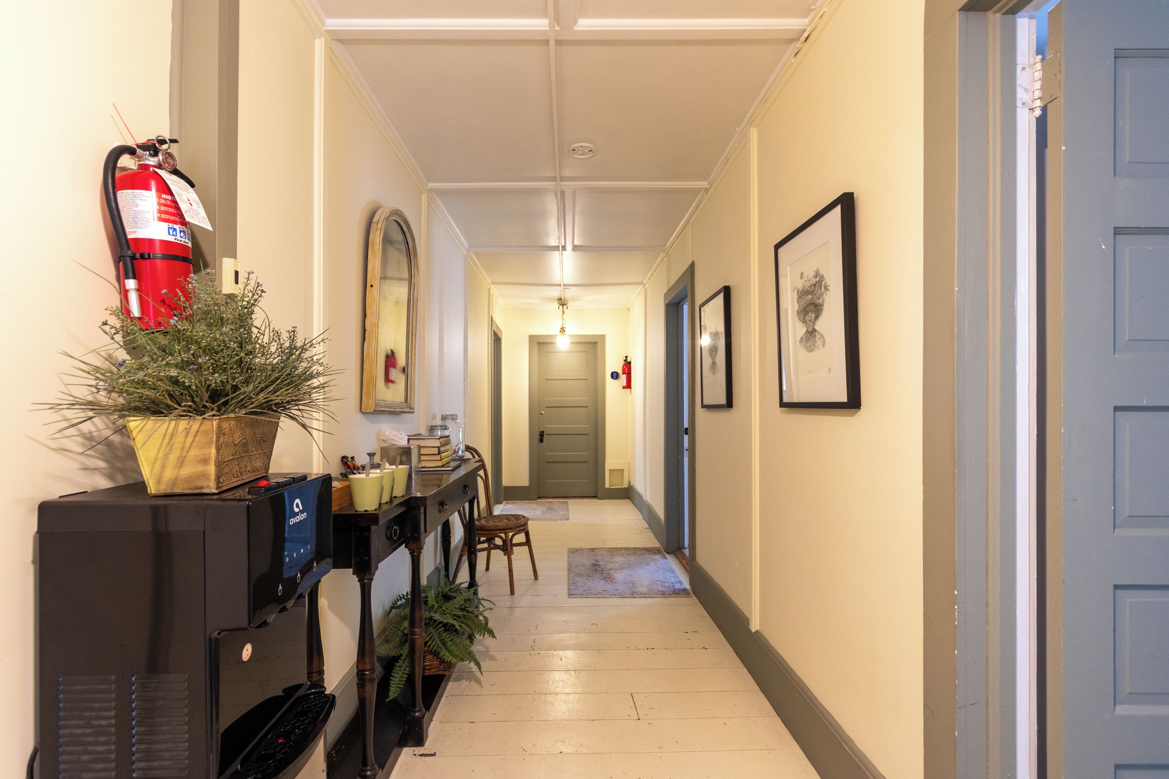 Un couloir avec un extincteur, une plante en pot, un bureau avec un ordinateur, des tableaux encadrés accrochés au mur et une porte au fond.