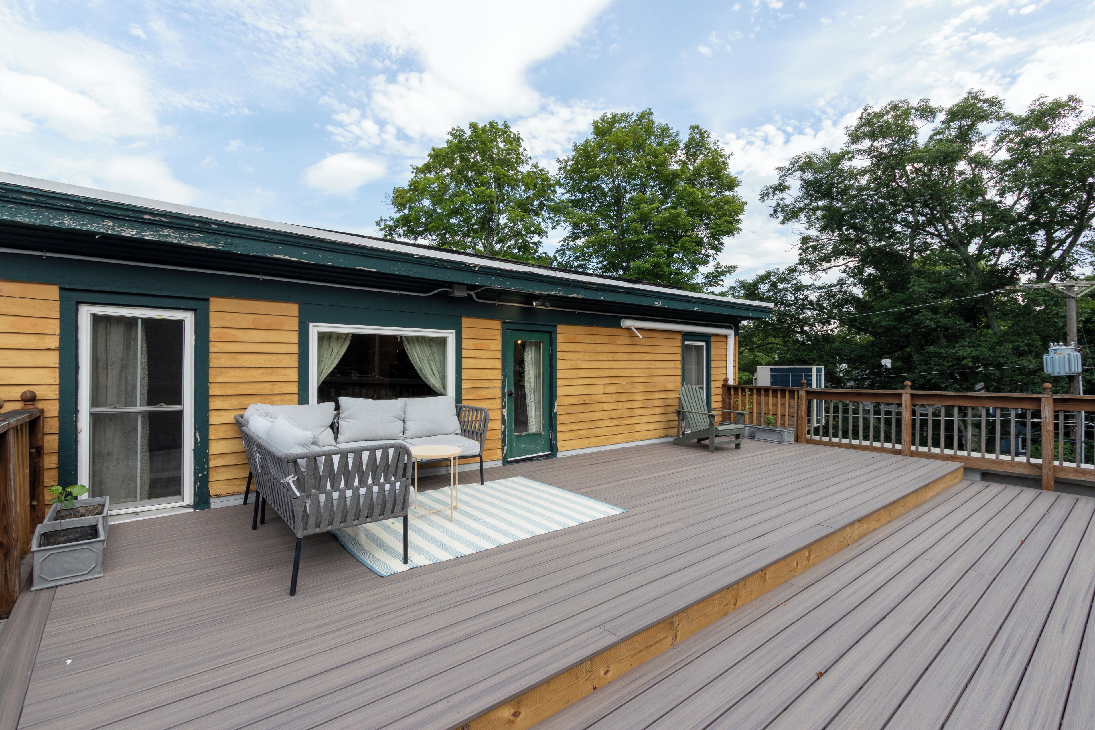 A wooden deck with a sofa and chairs, a covered patio area, and a view of trees.