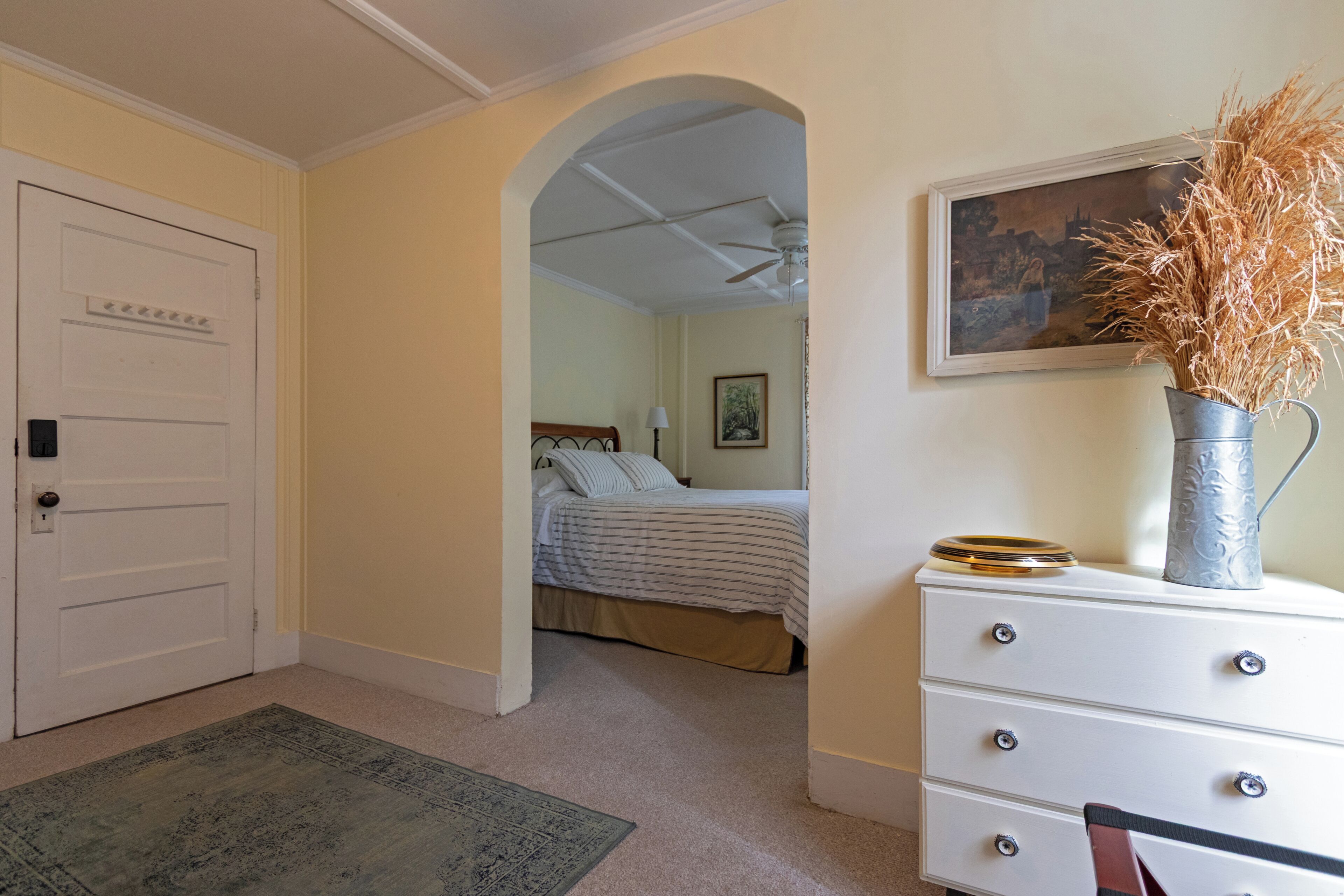 Une chambre à coucher avec un lit, une commode, un tableau et un vase contenant des plantes séchées.