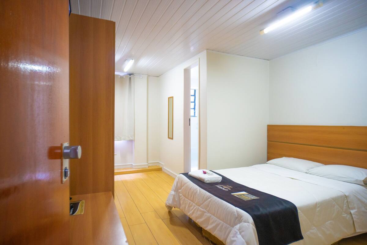 Una habitación de hotel con una puerta de madera, una cama con ropa blanca, un plaid oscuro y un pequeño cuadro en la pared.