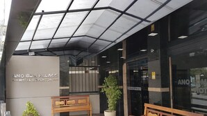 La entrada del Hotel Palacio Ano Bomi & Eventos, con un diseño moderno, cuenta con fachada de vidrio, bancos de madera y una planta en maceta.