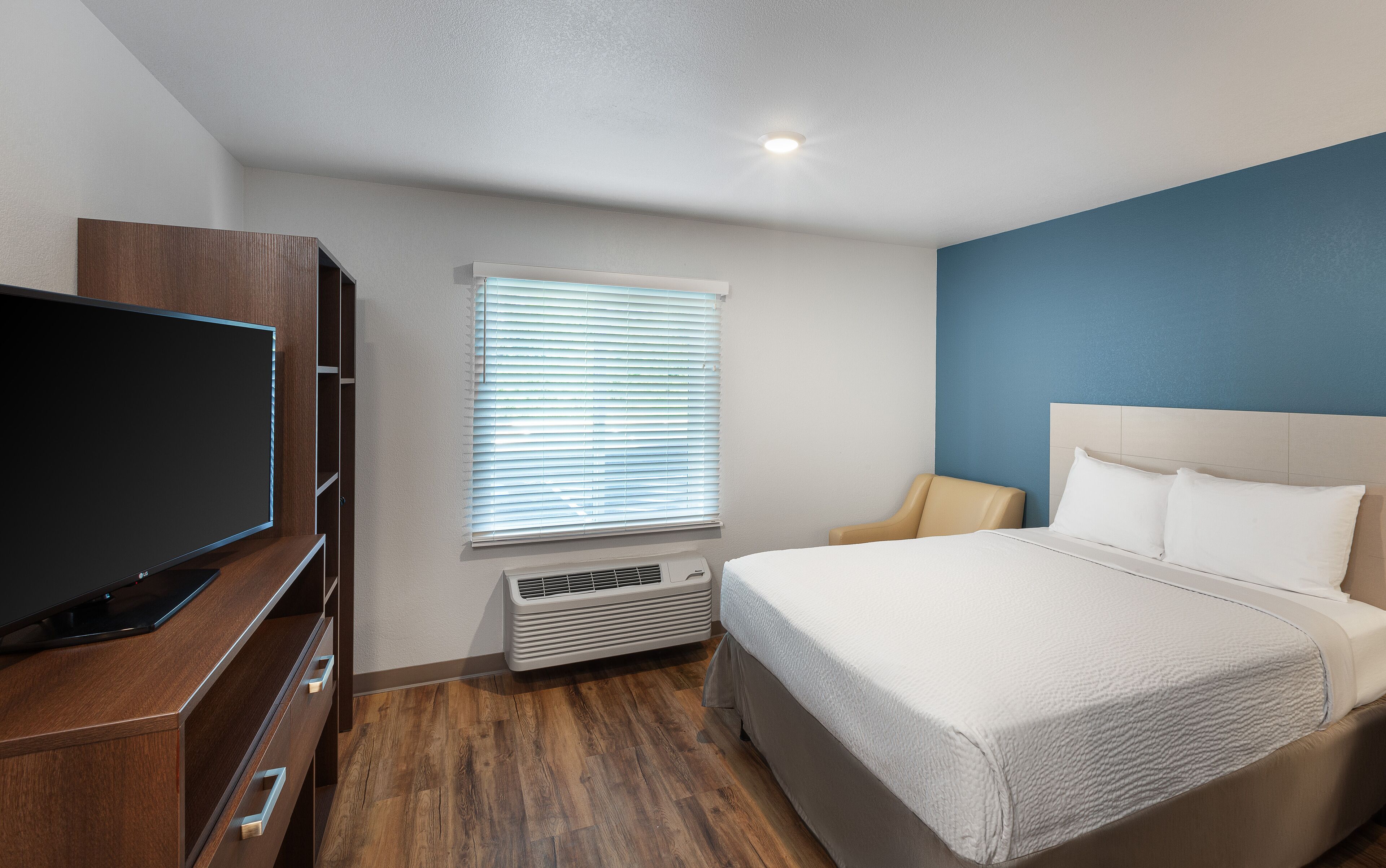 Een hotelkamer met een bed, een televisie, een raam met jaloezieën en een wandheater.