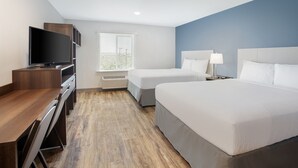 A hotel room with a wooden desk, a flat-screen TV, two beds, a window with blinds, and a nightstand with a lamp.