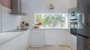 A modern kitchen with white cabinetry, a dishwasher, a stainless steel range hood, a built-in oven, and a large window offering a view of greenery.