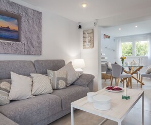 Ein modernes Wohnzimmer mit grauem Sofa, gemusterten Kissen, einem weißen Couchtisch und einem Essbereich mit Glastisch und Stühlen.