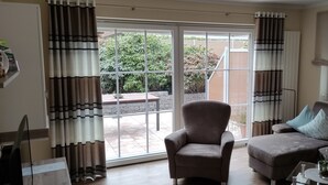 Un salon comprenant un canapé, un fauteuil et une table basse en verre. Une porte-fenêtre coulissante donne sur un patio doté d’un sol en briques et bordé de verdure.