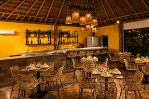 Un restaurante con sillas y mesas de mimbre, una zona de bar y un techo de paja.