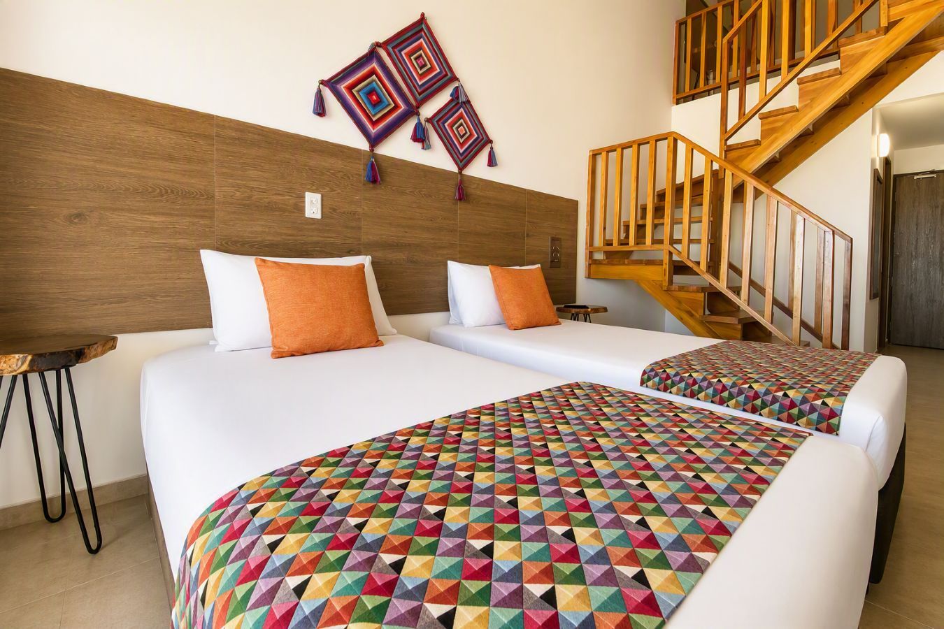 A hotel room with two beds, a wooden headboard, a staircase, and a colorful geometric-patterned bedspread.