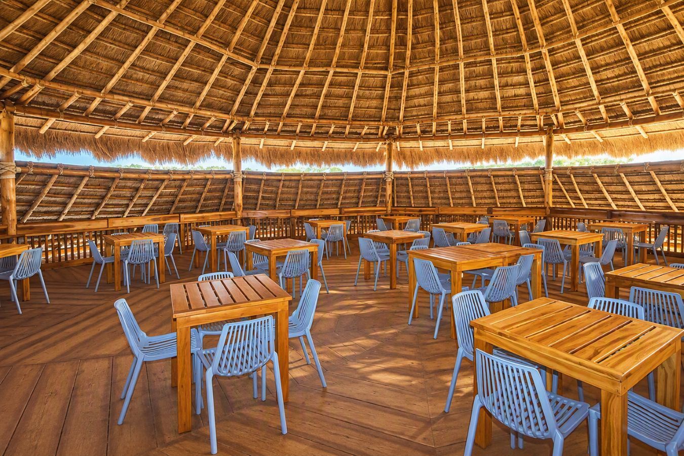 An open-air dining area with wooden tables and chairs, a thatched roof, and a wooden floor.