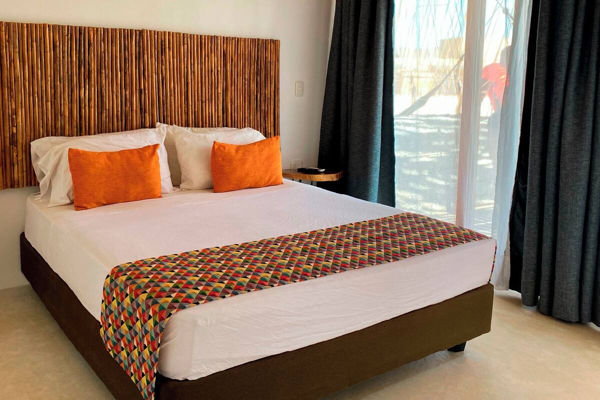 A hotel room with a large bed, a wooden headboard, two orange pillows, a colorful quilt, a small wooden side table, and a view of the outdoors through a window with curtains.