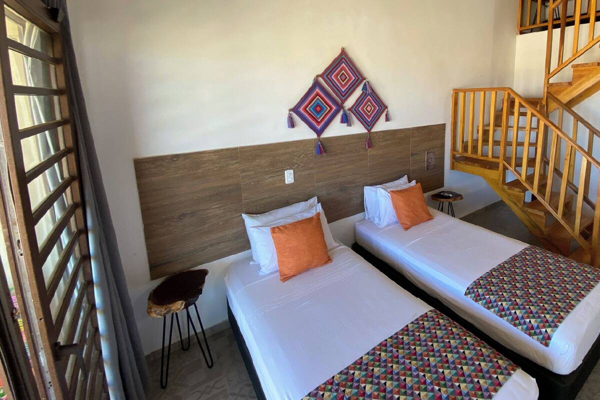 A hotel room with two beds, a wooden headboard, a small stool, a wall hanging, and a wooden staircase.