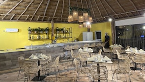 An outdoor dining area with wicker chairs and tables, a bar with a yellow wall and shelves, and a thatched roof ceiling. - Hotel Wayira Beach by On vacation (Manaure)