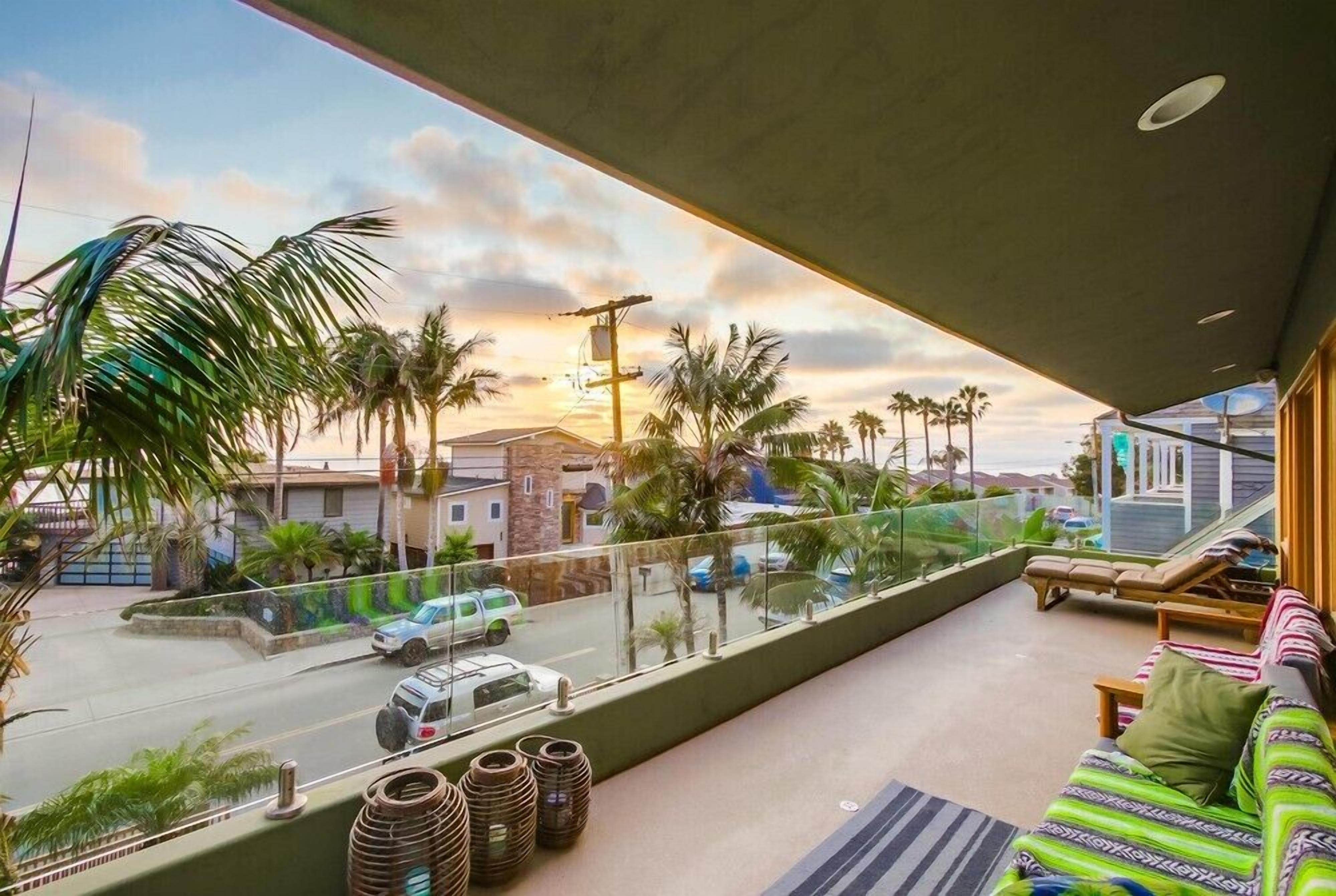 Un balcón con vista a una calle, palmeras y un atardecer.