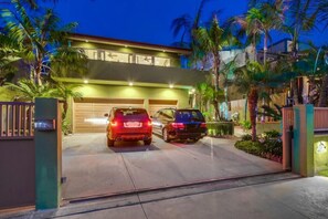 A modern two-story house with a garage, two cars parked in front, and a well-lit driveway surrounded by palm trees.
