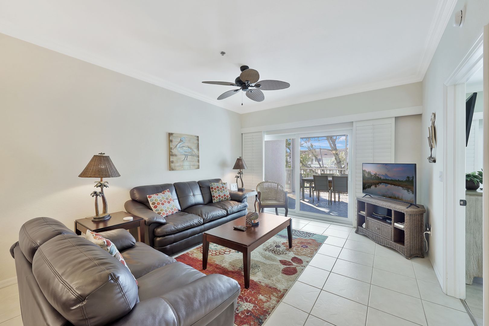 A living room with a sofa, coffee table, and TV stand. There is a ceiling fan, a painting, and a view of the outdoors through a glass door.