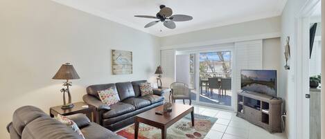 A living room with a sofa, coffee table, and TV stand. There is a ceiling fan, a painting, and a view of the outdoors through a glass door.