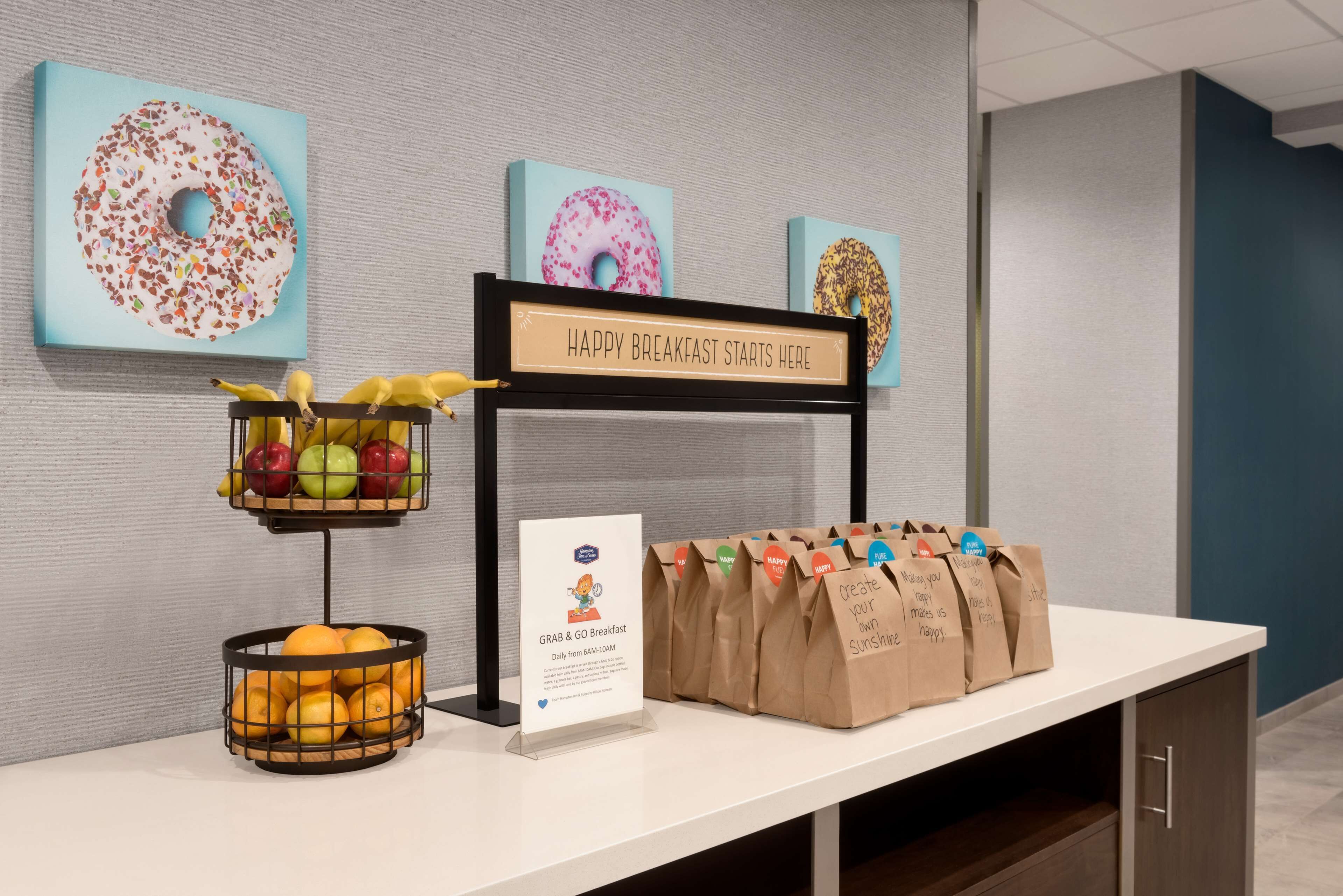 Una estación de desayuno con un cartel que dice “HAPPY BREAKFAST STARTS HERE”, una cesta de frutas y bolsas de papel con la leyenda “GRAB & GO BREAKFAST”.