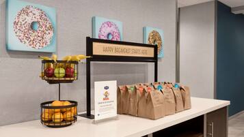 Una estación de desayuno con un cartel que dice “HAPPY BREAKFAST STARTS HERE”, una cesta de frutas y bolsas de papel con la leyenda “GRAB & GO BREAKFAST”.