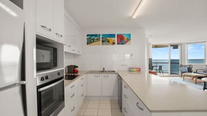Eine moderne Küche mit weißen Schränken, einem eingebauten Backofen und einer Mikrowelle. An der Wand hängen drei farbenfrohe Gemälde, und es gibt einen Balkon mit Meerblick.