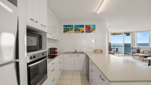 A modern kitchen with white cabinetry, a built-in oven, and a microwave. There are three colorful paintings on the wall, and a balcony with a view of the sea.