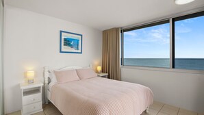 Ein Schlafzimmer mit Bett, Nachttischen, einer Lampe, einem gerahmten Bild und einem Fenster mit Meerblick.