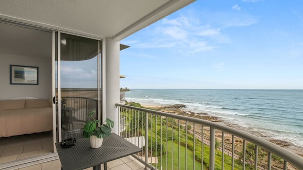 A balcony with a view of the ocean, a bed, a small table with a plant, and a chair.