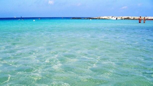 Agua turquesa cristalina en una playa con costa rocosa y algunas personas a lo lejos.