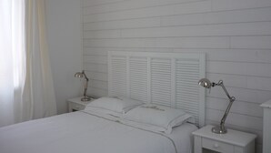 Ein Schlafzimmer in Weiß mit einem Bett, zwei Nachttischen, zwei Lampen und einem Korbwägelchen, das an der Decke hängt.