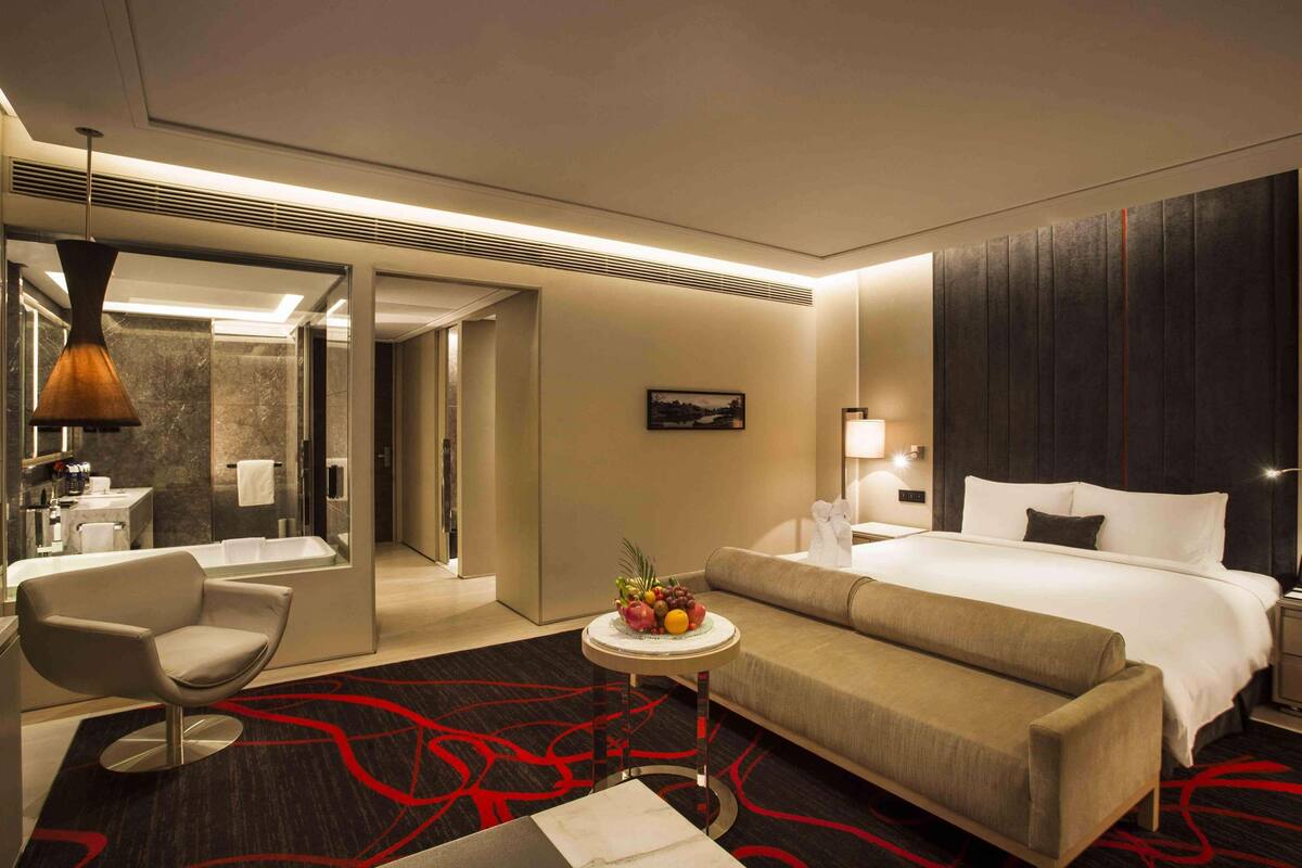 A modern hotel room with a large bed, a sofa, a round table with fruit, a chair, a bathroom, and a large mirror.
