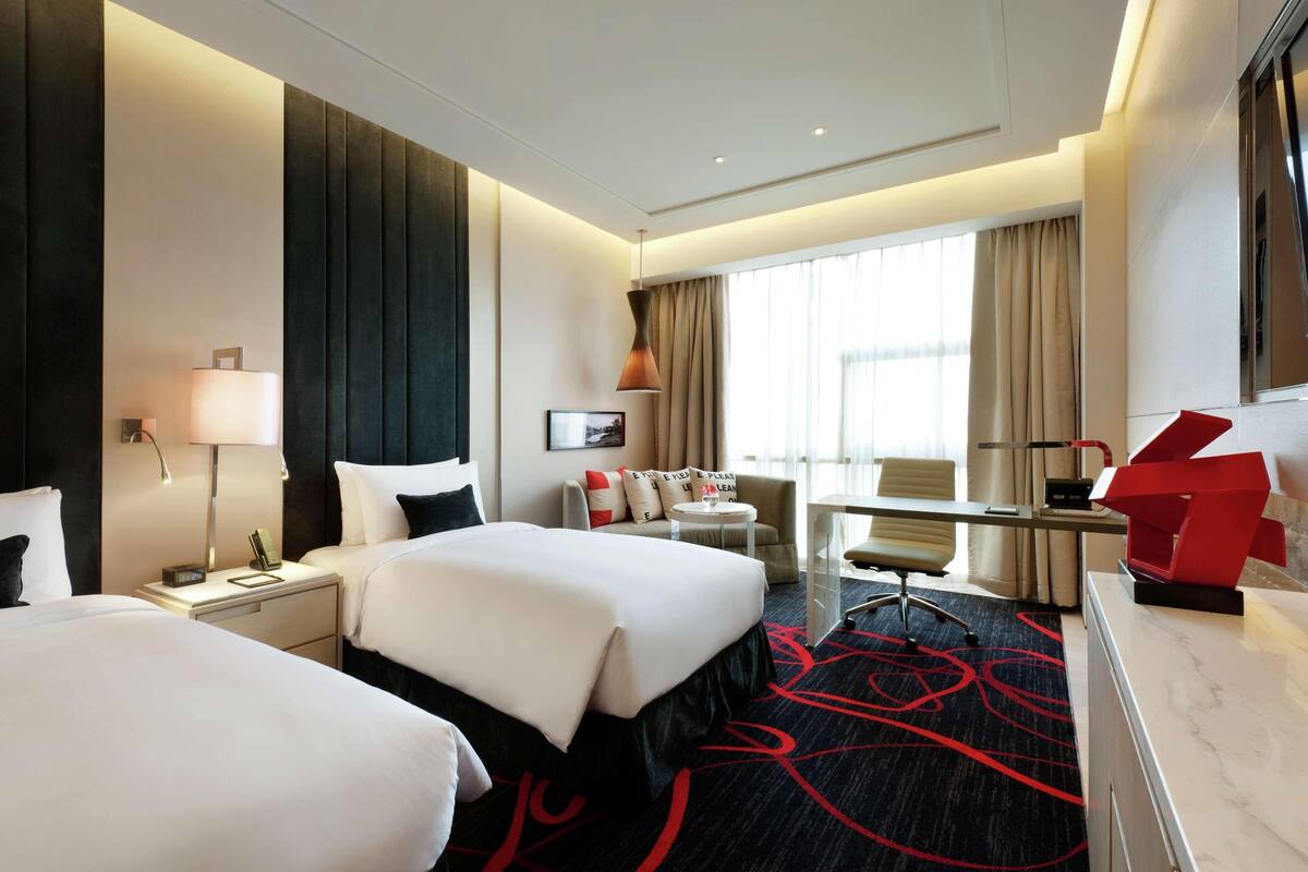 A hotel room with two beds, a desk, a chair, a lamp, a desk lamp, a picture frame, and a window with curtains.