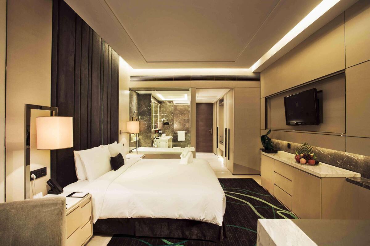 A modern hotel room with a large bed, bedside tables, a desk, a flat-screen TV, and a bathroom visible through a glass door.