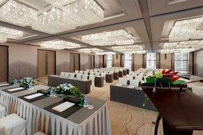 Une salle de conférence avec des rangées de chaises blanches, une longue table équipée de microphones et de grands lustres.