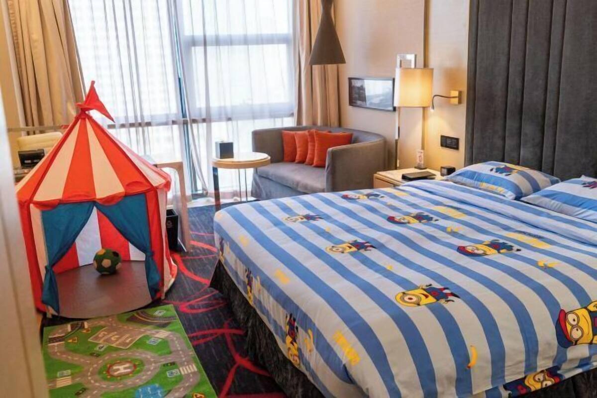 A hotel room with a bed, a small play tent, a round table, a sofa, and a lamp.