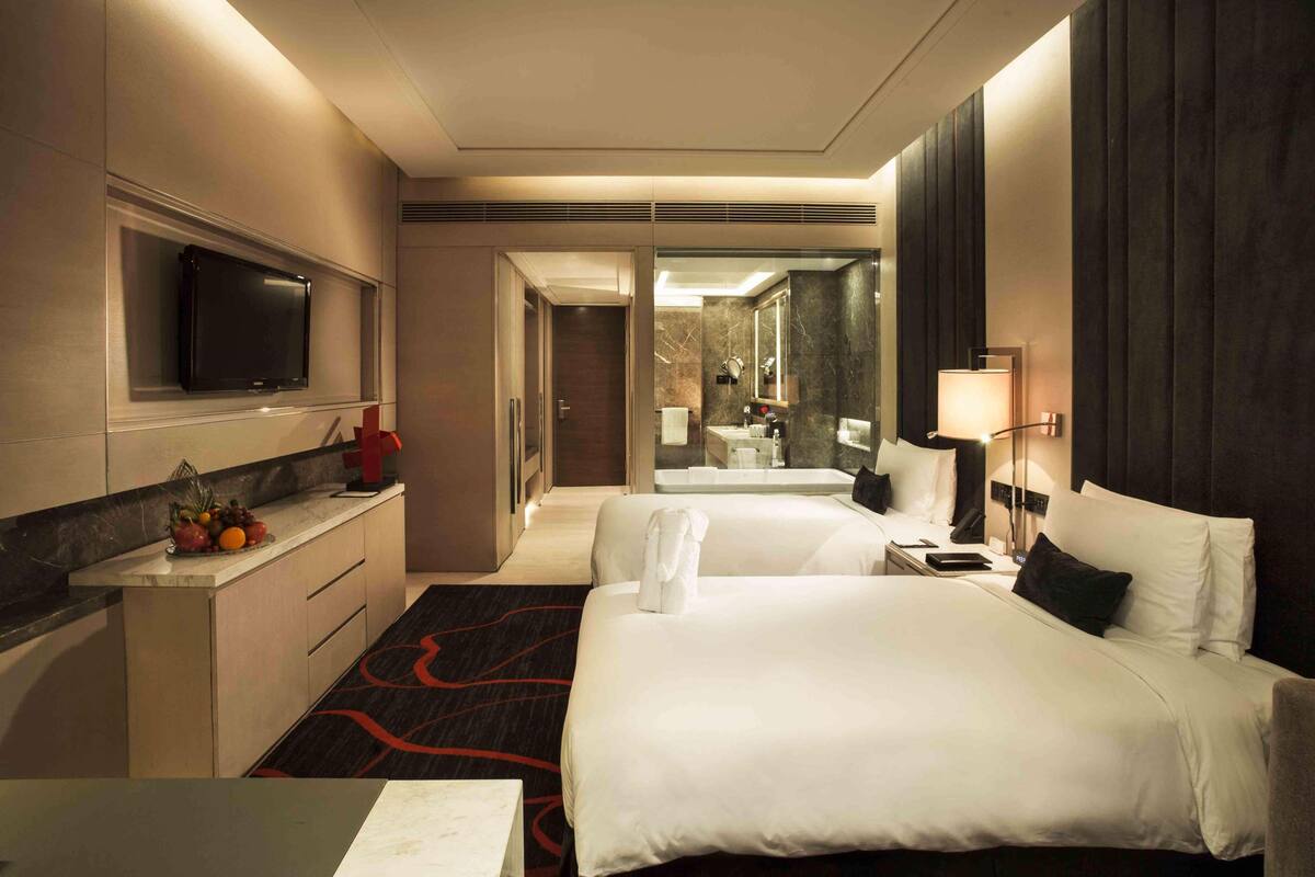 A hotel room with two beds, a TV mounted on the wall, a desk with a fruit bowl, and a bathroom visible through an open door.