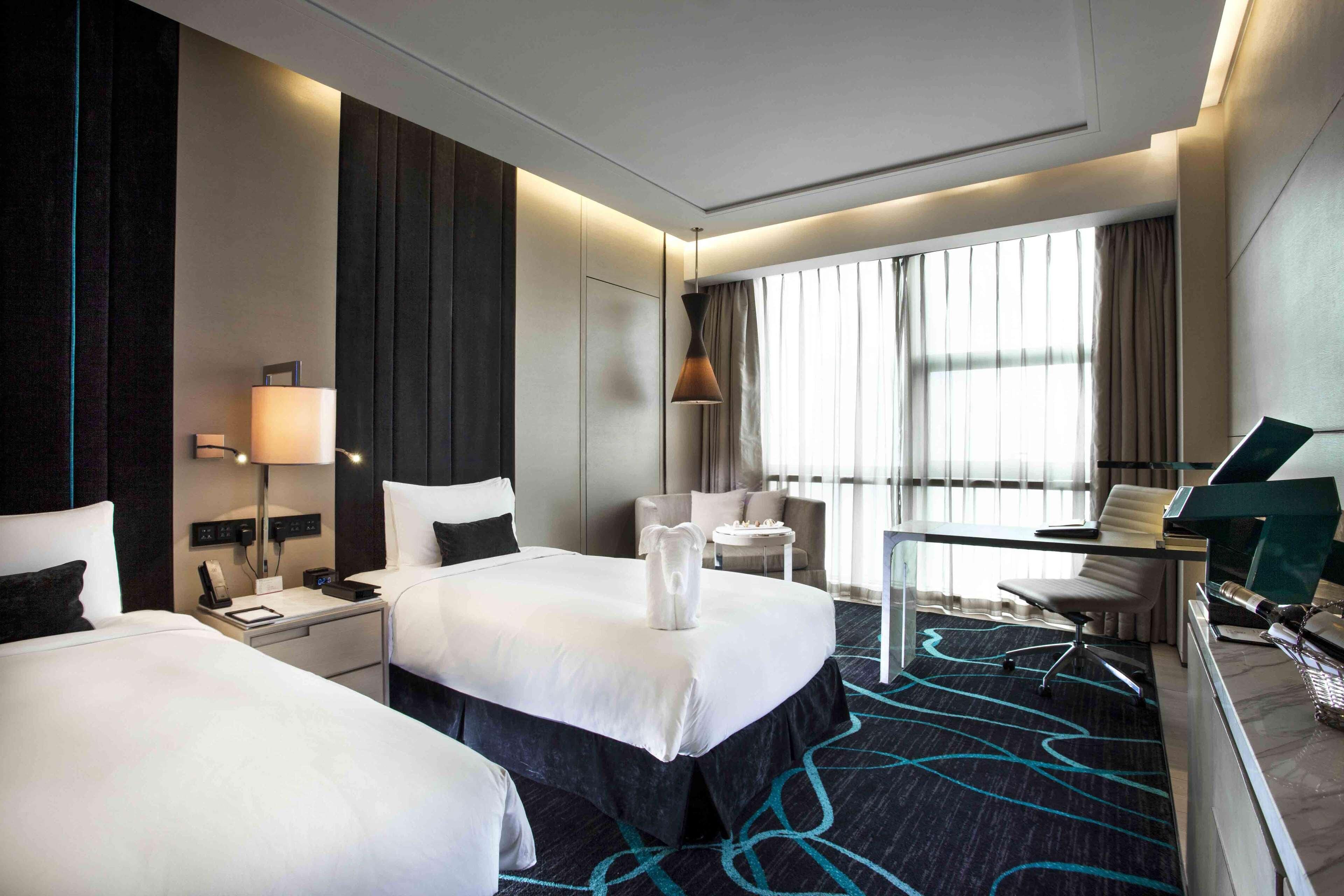 A hotel room with two beds, a desk, a chair, a lamp, and a large window with curtains.