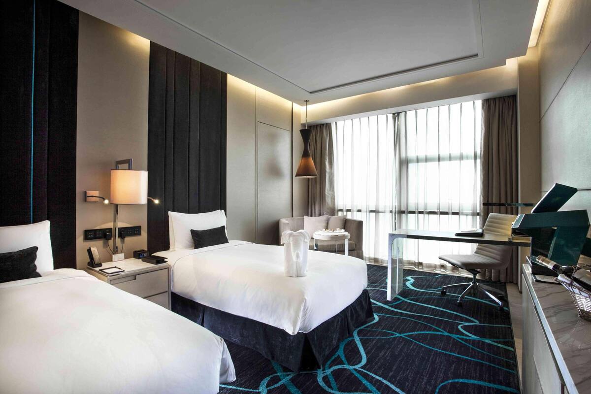 A hotel room with two beds, a desk, a chair, a lamp, and a large window with curtains.