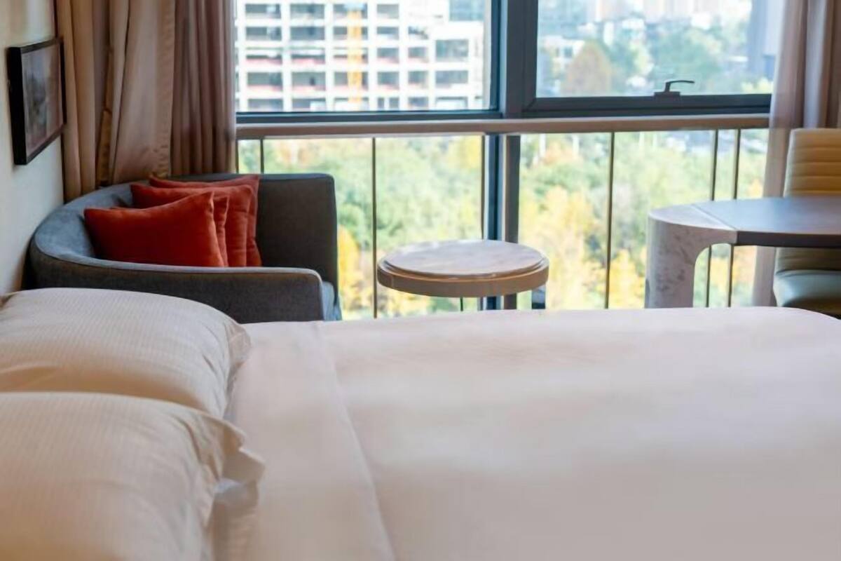 A hotel room with a bed, a small round table, a chair, and a view of the city through large windows.
