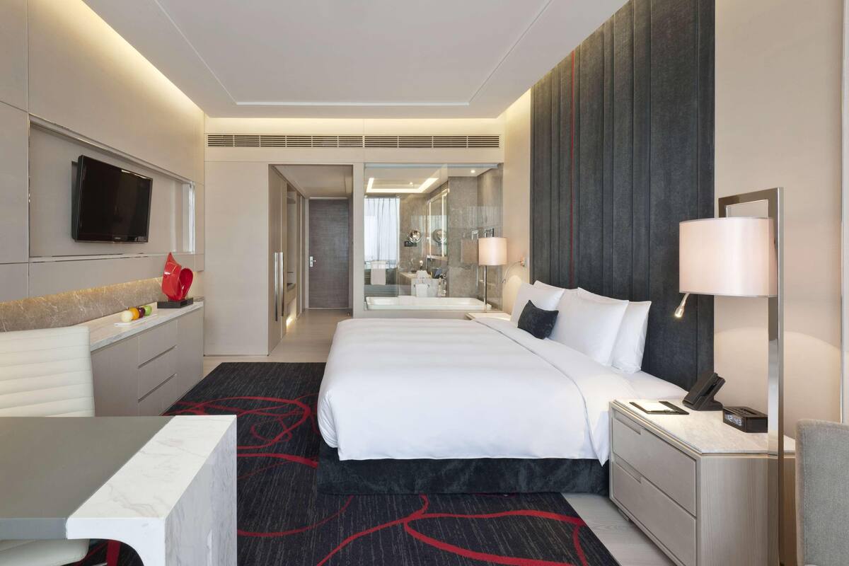 A modern hotel room with a large bed, bedside table, TV, and a bathroom visible through an open door.