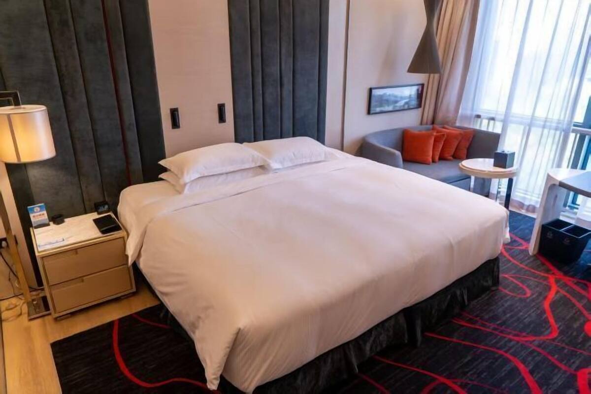 A hotel room with a large bed, a desk, a chair, and a small table with a lamp.