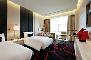A hotel room with two beds, a desk, a chair, a lamp, a desk lamp, a picture frame, and a window with curtains.