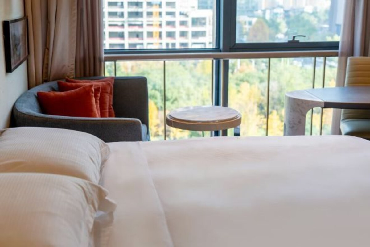 A hotel room with a bed, a small round table, a chair, and a view of the city through large windows.