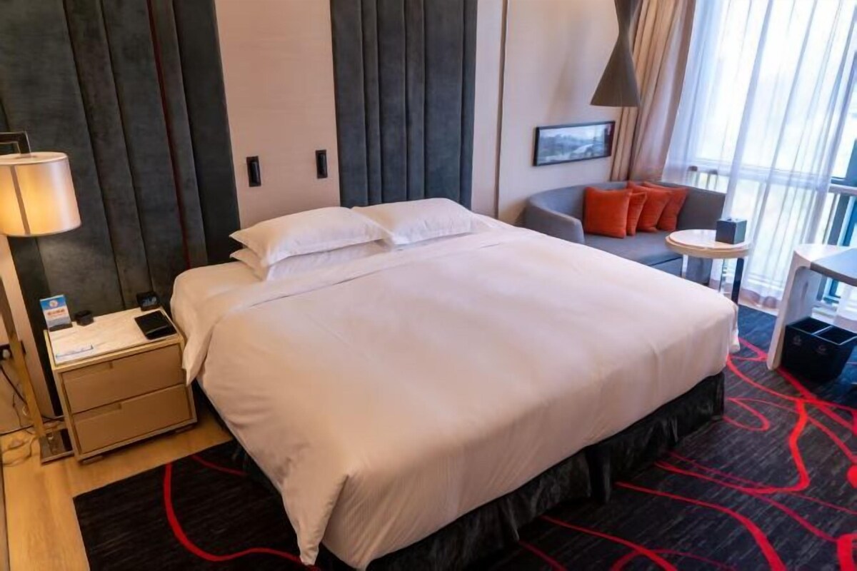 A hotel room with a large bed, a desk, a chair, and a small table with a lamp.