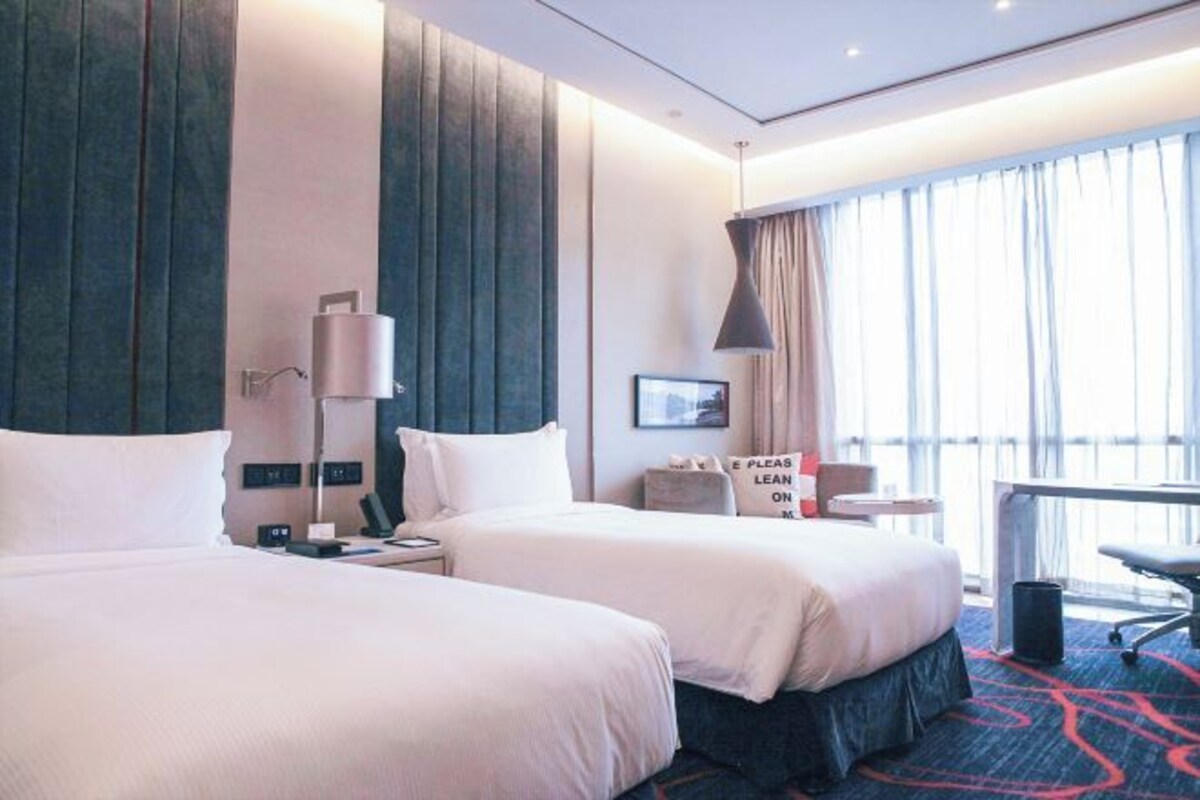 A hotel room with two beds, a desk, a chair, a lamp, and a large window with curtains.