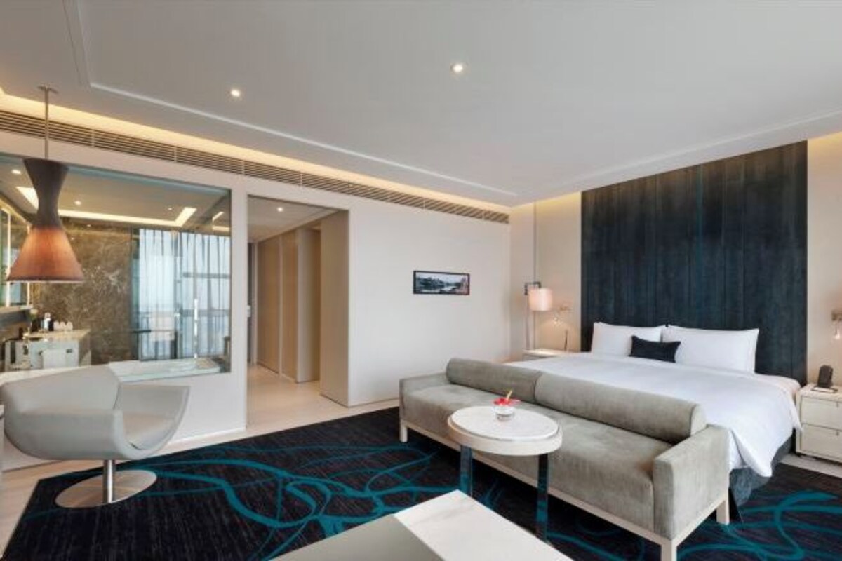 A modern hotel room with a large bed, a sofa, a small round table, and a chair. There is a bathroom visible through a glass door.