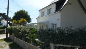 A two-story house with a white exterior, a visible chimney, and a small garden area with bushes and a tree.