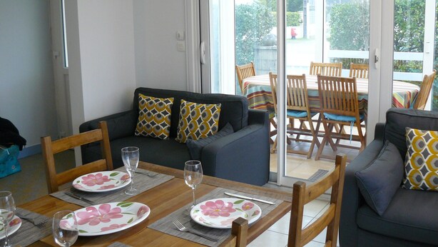 A dining area with a table set for two, a sofa with patterned cushions, and a view through a glass door to an outdoor patio.