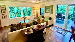 A spacious living room with a ceiling fan, a dining area with a table and chairs, and large windows offering a view of greenery.