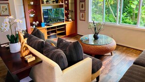 A living room with a sofa, a round glass table, a TV, and a window with a view of greenery.